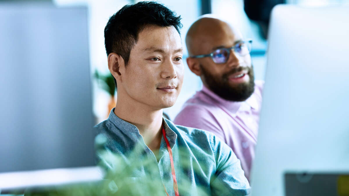Two male co-workers looking at computer screen and concentrating, mid adult Asian man wearing lanyard, freelance worker